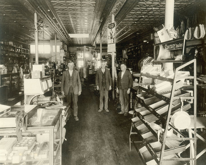 Historic interior of Nixa Hardware with three men, merchandise displays, tall shelving, and a patterned tin ceiling