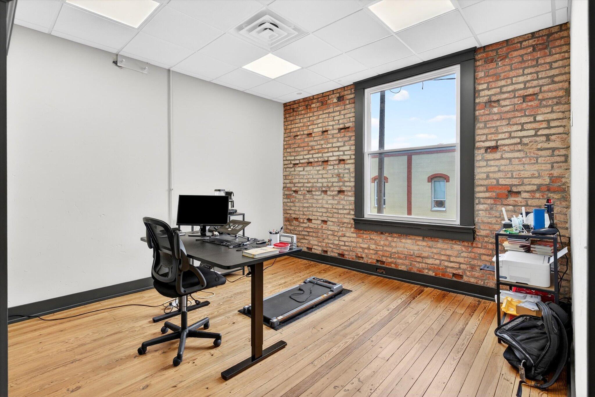 An existing private office with a large window, exposed brick, and wood floors